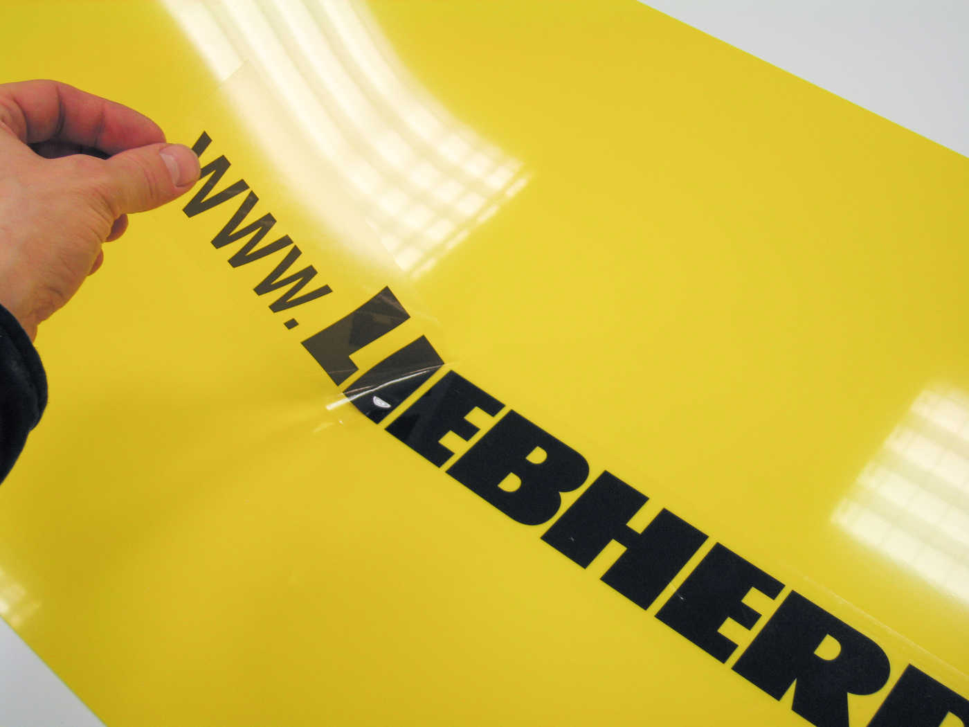 Gjennomsiktig selvklebende merke med Liebherr-logo monteres på gul bakplate.