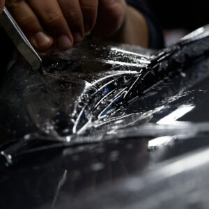 Nærbilde av en person som forsiktig skjærer til gjennomsiktig beskyttelsesfilm (PPF) på en våt biloverflate med en presisjonskniv.Paint protection film Gjøvik