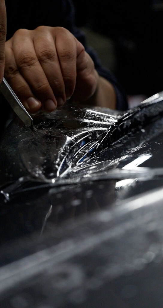 Nærbilde av en person som forsiktig skjærer til gjennomsiktig beskyttelsesfilm (PPF) på en våt biloverflate med en presisjonskniv.Paint protection film Gjøvik